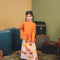Young girl in an orange top and patterned skirt standing among suitcases against a colorful wall.