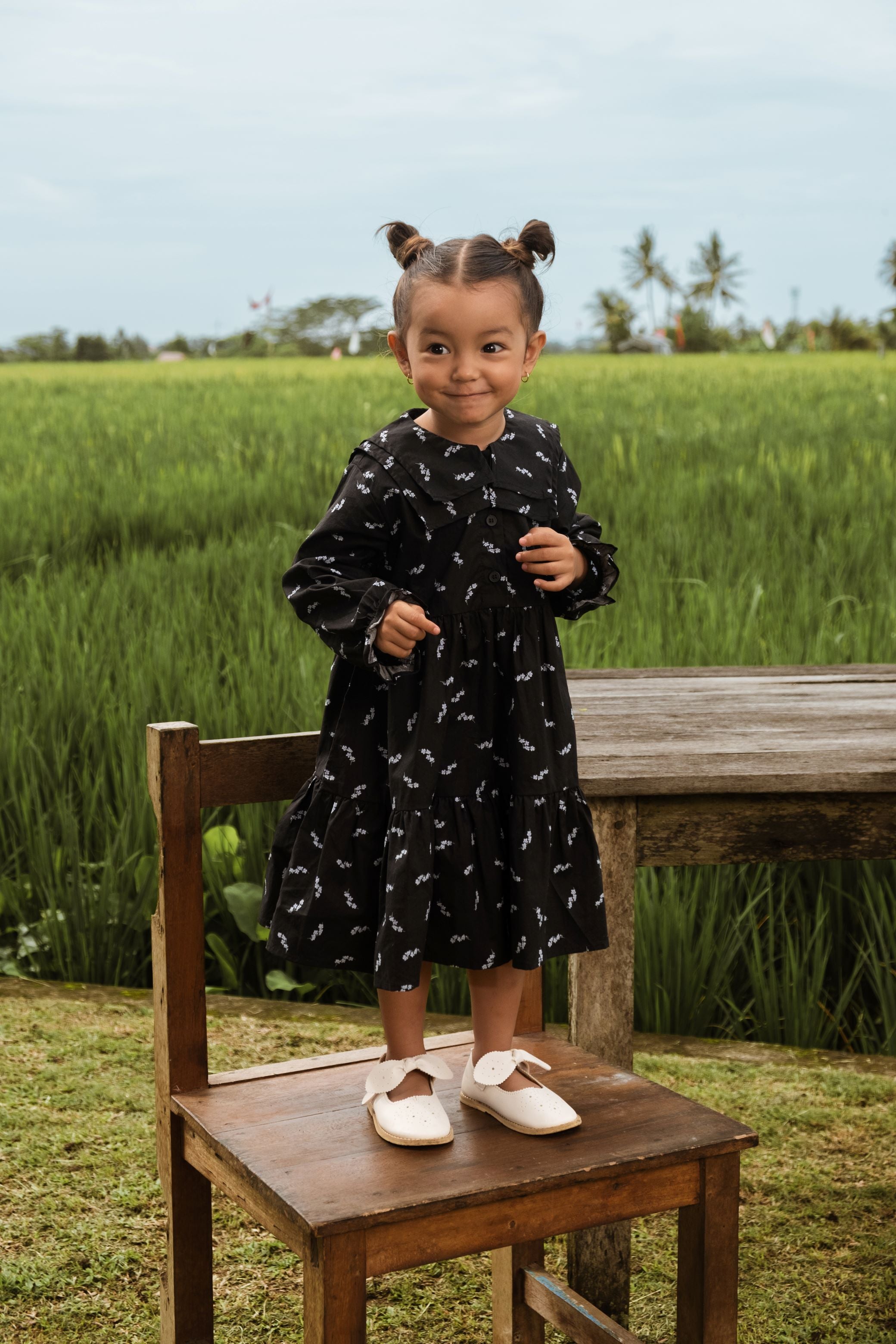 cheerful little girl donning melati print holiday dress, while standing on chair