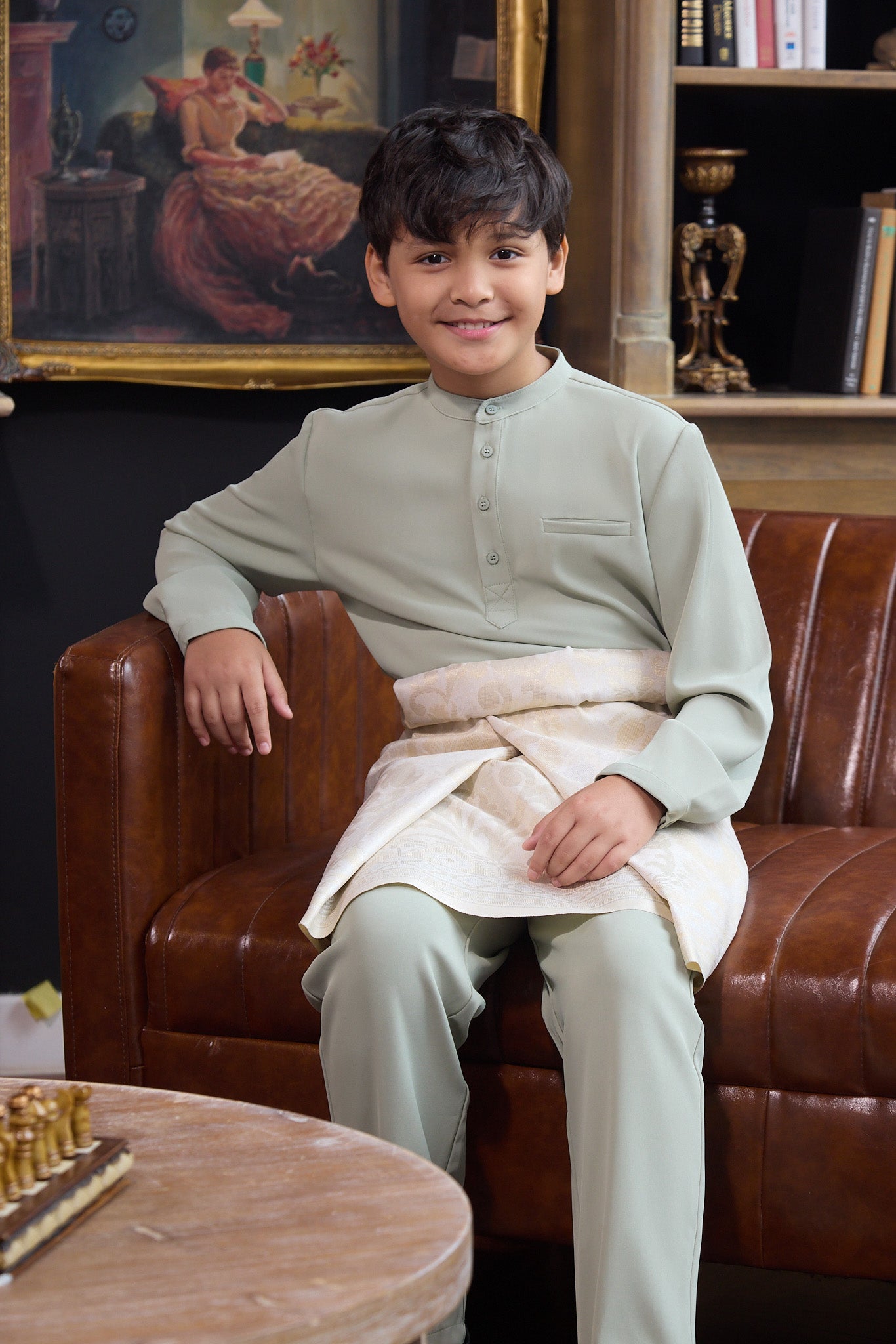 young boy smiling and sitting on chair wearing sage green boy baju melayu set